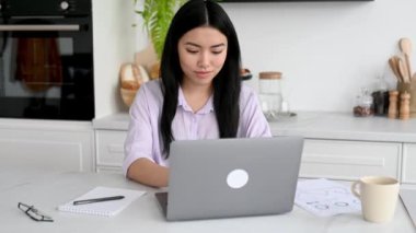 Uzaktan öğrenme, online ders. Odaklanmış zeki Çinli kız öğrenci, şık günlük giysiler içinde, uzaktan çalışıyor, dizüstü bilgisayar kullanıyor, çevrimiçi dersler sırasında not alıyor, evde oturuyor.