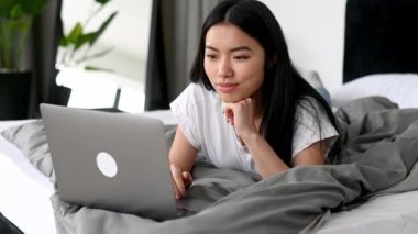 Güzel pozitif Asyalı esmer kız, gündelik giysiler içinde, evde yatakta yatıyor, dizüstü bilgisayar kullanıyor, sosyal ağlara takılıyor, arkadaşlarıyla sohbet ediyor, haber okuyor, gülümsüyor, evde dinleniyor.
