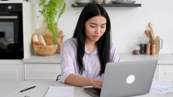 スマート素敵な正アジアのブルネットの女の子でシャツ デザイナー フリーランスまたは学生 ノートパソコンでキッチンに家に座って 仕事や研究をリモートで ノートを取り 笑顔優しい — ストック動画