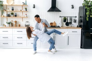 Neşeli, mutlu, birbirine aşık romantik çift, şık kıyafetler içinde, karışık ırk adamı ve beyaz kız, evde birlikte eğleniyorlar, mutfakta dans ediyorlar, gülümsüyorlar, eğleniyorlar.