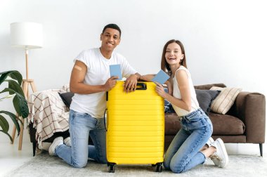Uzun zamandır beklenen tatil, seyahat. Mutlu çok ırklı bir çift, aşıları var ve seyahate çıkıyorlar, oturma odasında, sarı bavulun yanında oturuyorlar, pasaportlarını tutuyorlar, kameraya bakıyorlar, gülümsüyorlar.