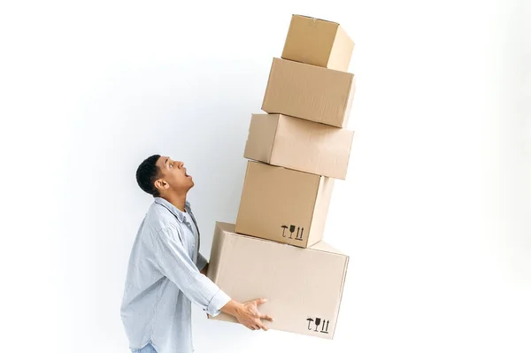 Delivery concept. Frightened mixed race delivery man, looking excitedly at a mountain of cardboard falling boxes he holds in his hands, with breakable goods inside, standing isolated white background