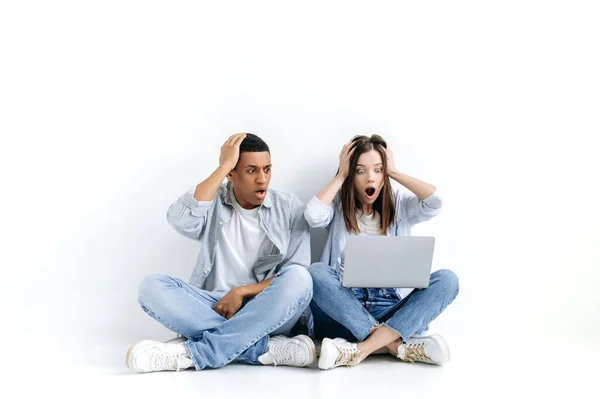 Shocked discouraged young hispanic man and caucasian woman, stylishly dressed, sitting on floor over isolated white background with laptop, looking at a screen in surprise, saw unexpected news