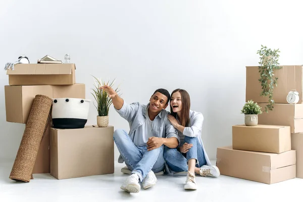 Happy young multiracial couple, caucasian woman and hispanic guy, are sitting on the floor between boxes of stuff in their new home or rental apartment, planning their house future design, smile