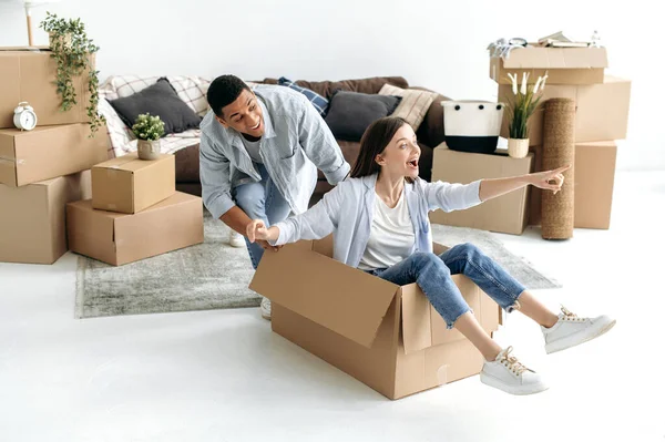 多种族快乐的夫妻 乐于购买新的房子 拥有自己的房子 玩纸板箱 女孩坐在里面 男人推着 他们在新公寓里玩得很开心 — 图库照片