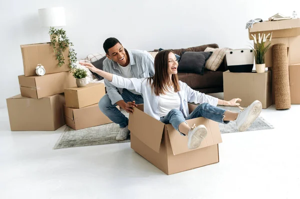 一对兴高采烈的恋爱中的夫妇 对自己的新房感到满意 他们正在玩纸板箱 女孩坐在里面 男人推着纸箱 他们一起在新公寓里玩乐 — 图库照片