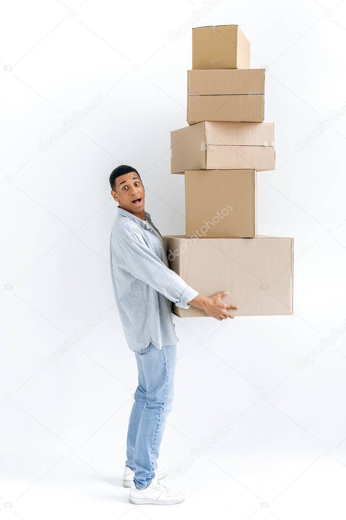 Excited mixed race guy in casual stylish clothes, delivery man, standing over isolated white background, holding many cardboard boxes, delivering parcels, looking in amazement at camera, delivery