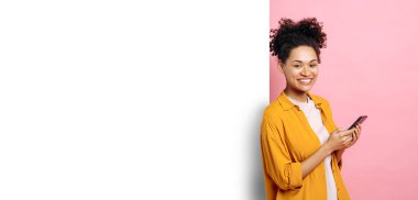 Afro-Amerikan kadınının panoramik fotoğrafı, akıllı telefon kullanıyor, sosyal medyada geziniyor, arkadaşlarıyla sohbet ediyor, pembe arka planda beyaz model pankartın yanında duruyor, kameraya bakıyor, gülümsüyor.