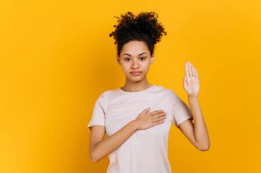 Sadece gerçeği. Dürüst, kıvırcık saçlı, sıradan tişörtlü Afro-Amerikan kadını, elini göğsüne koyuyor ve avuç içi kaldırıyor, söz veriyor, yemin ediyor, izole edilmiş turuncu arka planda duruyor.