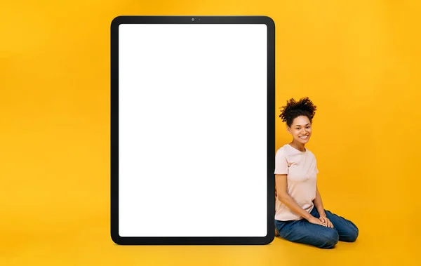 Full length photo of excited African American girl sits near an enlarged tablet with blank white mock-up screen for presentation or advertising, looks at camera, smiles, orange isolated background