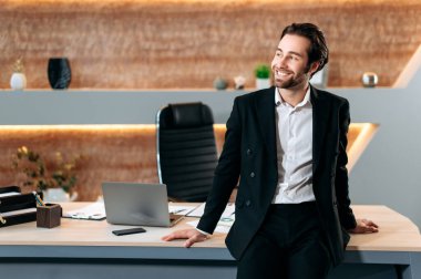 Pozitif profesyonel başarılı Kafkasyalı yönetici, iş adamı, şirketin CEO 'su, şık bir iş elbisesi içinde, modern bir ofiste masasının yanında duruyor, yana bakıyor, mutlu bir şekilde gülümsüyor.