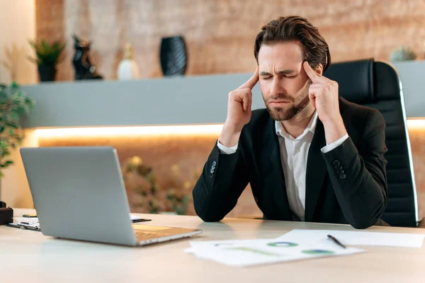 Fatigue caucasian businessman, office employee, sits at a workplace in a modern office, touch his temples, massage head, feel stressed, overworked, headache, migraine, need rest and coffee brak