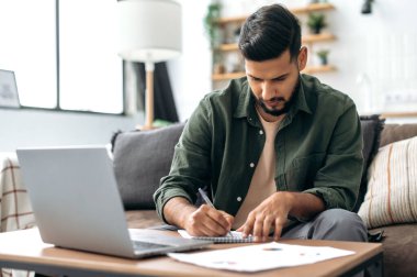 Odaklanmış akıllı Hintli ya da Arap adam, öğrenci ya da serbest çalışan, oturma odasında kanepede oturan, dizüstü bilgisayar kullanan, çalışan ya da uzaktan çalışan, webinar dinleyen, notlar alan, ödev alan,