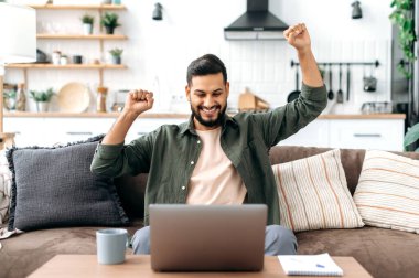 Arap ve Hint uyruklu mutlu bir adam, oturma odasında bir koltukta oturur, dizüstü bilgisayara bakar, elleriyle el kol hareketi yapar, başarıyı kutlar, zafer, büyük kazanç, iyi haberler alır, gülümser,
