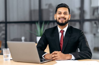 Pozitif genç, kendinden emin Arap ya da Hintli bir işadamının portresi, takım elbiseli başarılı bir girişimci, modern yaratıcı ofiste dizüstü bilgisayarla çalışma masasında oturuyor, kameraya bakıyor, dostça gülümsüyor.