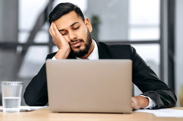 Sleepy arabian or indian business man in a suit, sits at workplace in the office with a laptop, put his head on his hand, fell asleep during work, tired, overworked, experiences chronic lack of sleep