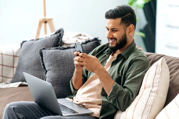Happy calm arabian or indian guy in casual clothes, spending time at home, sits on the sofa in the living room, using a smartphone, chatting with friends, browsing the internet, social networks, smile