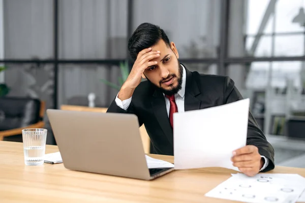 Stressed tired indian or arabian business man, sitting upset at a work desk with laptop in modern office, works with documents, makes a financial report, analyzes the result of work, feels overworked