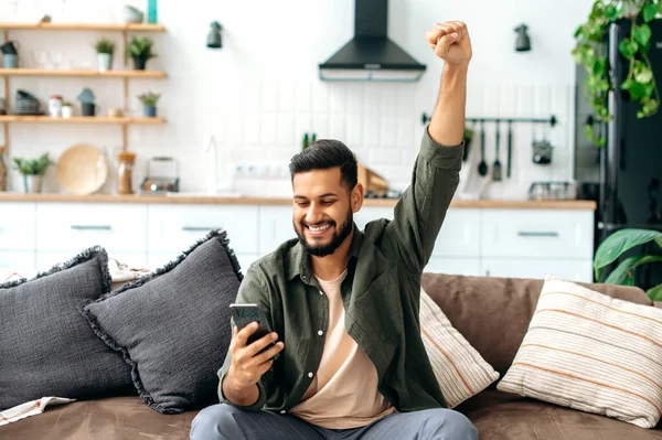Win, success. Indian cheerful happy young indian or arabian man in casual wear, sit at home in living room on comfortable sofa, using smart phone, read good news, gesturing with hand, smiling joyfully