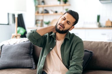 Hintli ya da Arap genç adam, rahat bir oturma odasında kanepede oturuyor, boynuna masaj yapıyor, yoğun bir iş gününden sonra yorgun, eklem problemleri çekiyor, boynunda sinir sıkışması, dinlenmeye ihtiyacı var.