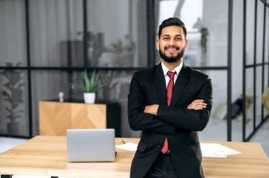Yakışıklı, kendine güvenen başarılı Hintli ya da Arap iş adamı takım elbiseli, erkek girişimci, masanın yanındaki modern ofiste kollarını kavuşturmuş, kameraya bakıyor, dostça gülümsüyor.