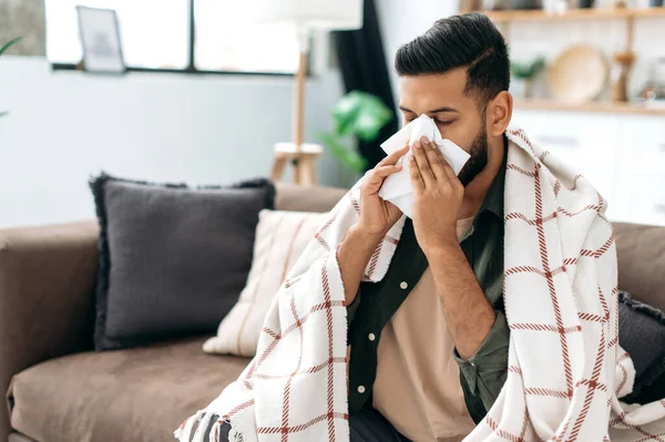 Sneezing, runny nose, cough. Ill arabian or indian young man, sits on the couch at home under a plaid, sneezing in a napkin, feels weak, unwell, needs treatment and medicines