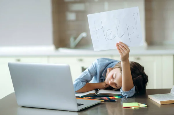 School online learning. Schoolgirl shows the inscription on the sheet HELP, while lying on the table while studying online using a laptop, feels tired of distance learning, overworked