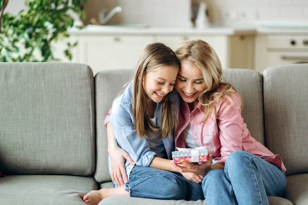 Happy mother and daughter, sitting together at home on the sofa in the living room, hugging, holding a small box with a gift in their hands, looking forward to seeing whats inside, smiling, rejoicing