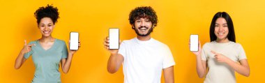 Panoramic photo of three multiracial people, indian guy, afro american and chinese girls showing smartphones with blank white mockup screens, looking at camera, smiling, isolated orange background