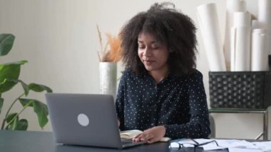 Dost canlısı, özgüvenli genç Afrikalı Amerikalı kadın serbest çalışan, öğrenci, menajer, yaratıcı ofiste ya da evde dizüstü bilgisayarla oturan, video görüşmesi yapan, konferans veren, not defterine notlar alan, gülümseyen