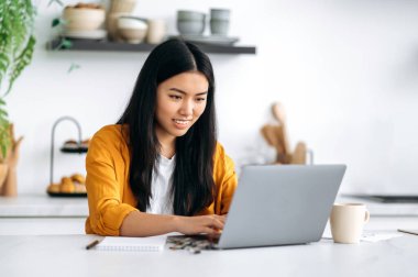 Positive friendly smiling Chinese girl, freelancer, programmer, manager, sitting at the table, working from home, using a laptop, working on a project with concentration, thinking over a strategy