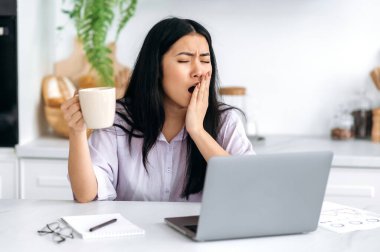 Sleepy Asian pretty girl working remotely, content manager, freelancer, student, sitting at the table in the kitchen with laptop, starting the working day, drinking coffee, yawning, unable to wake up