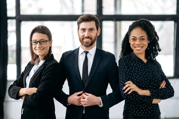 Portrait of successful group of multiracial business people in a modern office, looking at camera, smiling. Happy proud office employees, a man and two women, standing in meeting room of their office