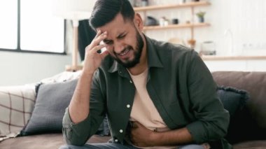Hint ve Arap kökenli üzgün bir adam, oturma odasında bir kanepeye oturuyor, ellerini karnının üstüne koyuyor, karnındaki ağrıdan dolayı yüzünü buruşturuyor, zehirlenme, spazm, mide sorunları yaşıyor.