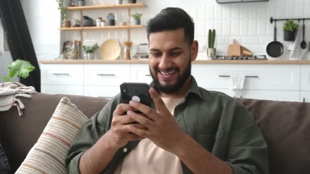 Positive Indian Arabian Guy Using His Smartphone While Sitting Sofa — Vídeo de Stock