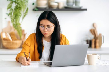 Çevrimiçi eğitim, e-öğrenme kavramı. Zeki, pozitif Asyalı, gözlüklü, başarılı bir öğrenci, bilgisayarını kullanarak evde çalışıyor, internetten web sitesi dinliyor, ders alıyor, notlar alıyor, arkadaşça gülümsüyor.