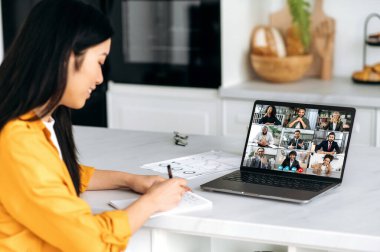 Çevrimiçi görüşmeler. Dizüstü bilgisayar başında çok ırklı bir grup insan ve mutfakta oturan Asyalı bir kadın bir video konferansında konuşuyorlar, bir proje stratejisi geliştiriyorlar, kız notlar alıyor.