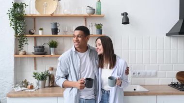 Mutlu çok ırklı romantik çift, yeni evliler, şık bir şekilde giyinmiş, mutfakta ayakta duruyor, ellerinde kahve ya da çay bardaklarını tutuyor, başlarını çeviriyor, hayal kuruyor, gülümsüyor, birlikte sabah vakitlerini geçiriyorlar.