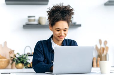 Oldukça başarılı genç Afrikalı Amerikalı kadın, serbest çalışan, bilişim uzmanı, mutfakta oturmuş, iş için dizüstü bilgisayar kullanıyor, iş arkadaşlarıyla mesajlaşıyor, bir proje üzerinde çalışıyor, mutlu bir şekilde gülümsüyor.