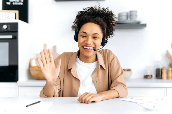 Positive confident curly-haired young african american woman with headset, call center worker, consultant, talks via video call, female mentor recording webinar, lecture, gesturing with hands, smiling