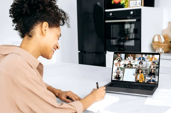 Video conference with teacher and students. Clever african american female student watching online lecture. Young woman takes notes, listening webinar from female teacher on video call