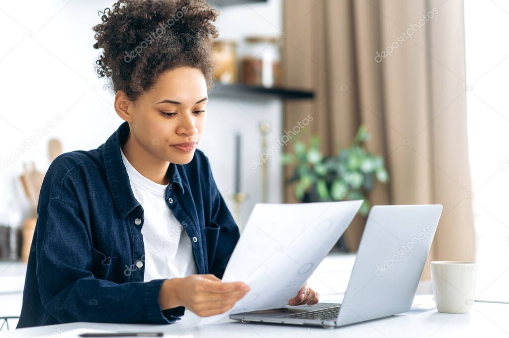 Concentrated young african american curly haired woman, freelancer, auditor, working from home, sitting in the kitchen, using a laptop, focused studying financial documents, graphs, reports