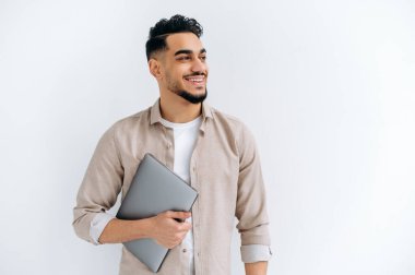 Cheerful arabian or indian young man with beard, wearing casual shirt, standing over isolated white background, holding laptop, looking to the side, smiling joyfully, dreaming, thinking