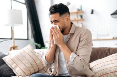 The concept of illness, runny nose. Unhealthy sad arabian or indian man sits at home in the living room on the couch, feels unwell, suffers from a runny nose, sneezing, uses napkins, needs treatment