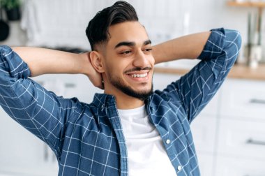 Mutlu, rahat bir Arap ya da Hintli adamın yakın plan fotoğrafı, serbest çalışan, mutfakta oturan, işe ara veren, ellerini başının arkasına koyan, rüya gören, gülümseyen