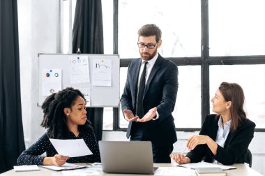 Takım çalışması konsepti. Takım elbiseli beyaz bir erkek patron, kadın çok ırklı meslektaşlarıyla iş görüşmesi yapıyor, yaratıcı modern bir ofiste, yeni bir iş projesini tartışıyor, stratejisini tartışıyor.
