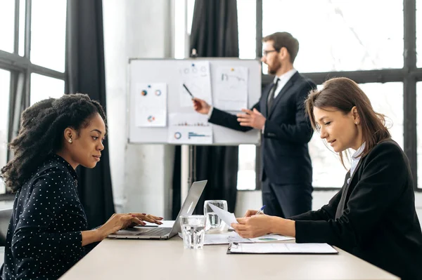 Concentrated clever female colleagues, an african american and a caucasian women are sitting at a table in a modern office, working on a project, develop new start-up, analizyng risks