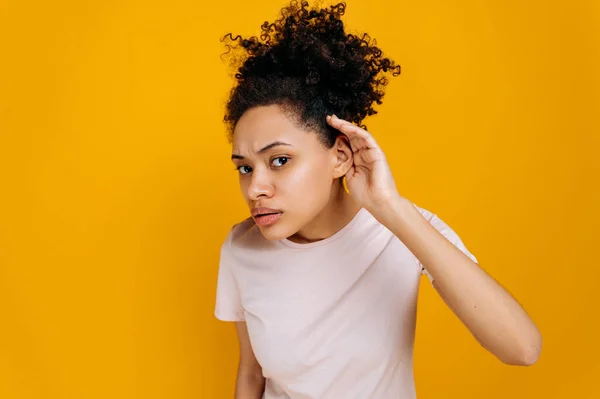 Deafness, gossip concept. African american young curly haired woman, wearing basic t-shirt with hand near ear, concentrated listening rumor, hearing gossip, standing on isolated orange background