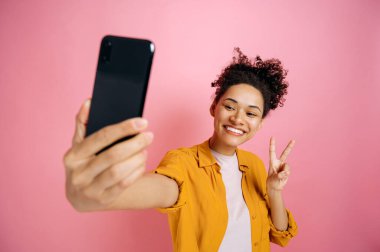 Happy positive african american excited girl, blogger, in casual stylish wear, doing selfie shot on cellphone and smiles into the camera, shows peace symbol, posing on isolated pink background, smiles