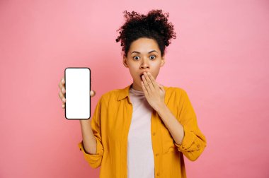 Confused shocked african american curly young woman in casual wear, showing smartphone with blank white mockup screen, standing on isolated pink background, looks surprised at camera, cover her mouth